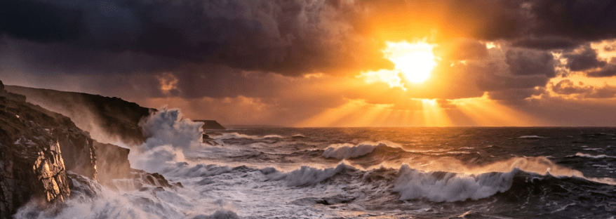 Sunrise illuminating dark clouds and powerful waves crashing on rocky shore
